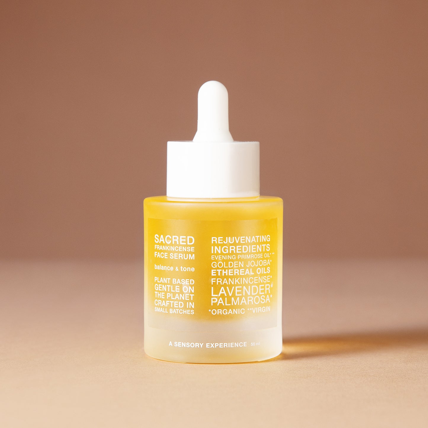 A 50 ml bottle of DEAR BOTANICS Artisanal Skincare face serum with a white dropper cap and a frosted glass bottle showing a golden-yellow liquid inside, set against a warm beige background.