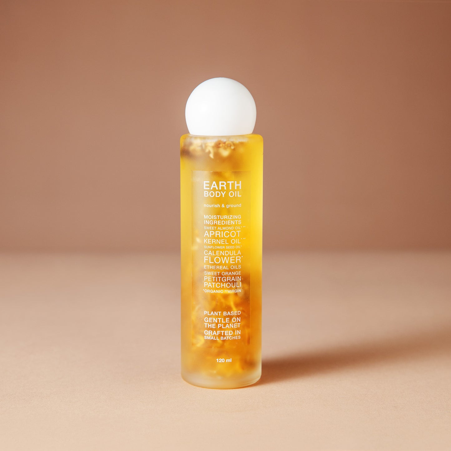 A 120 ml bottle of DEAR BOTANICS essential body oil, featuring a translucent yellow body filled with visible botanical pieces and a white spherical cap, set against a warm beige background.
