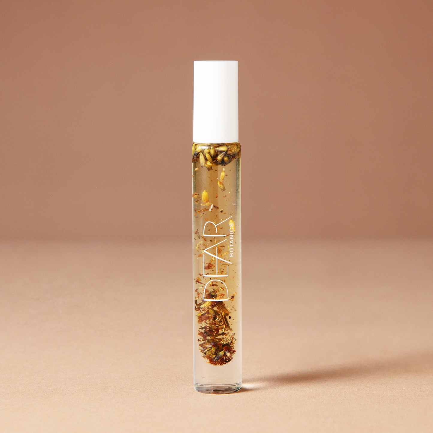A 10 milliliter clear cylindrical roller bottle filled with pale yellow perfume oil infused with dried flowers featuring a white cap and white vertical text that reads 'DEAR BOTANICS essential oil', set against a solid warm beige background.