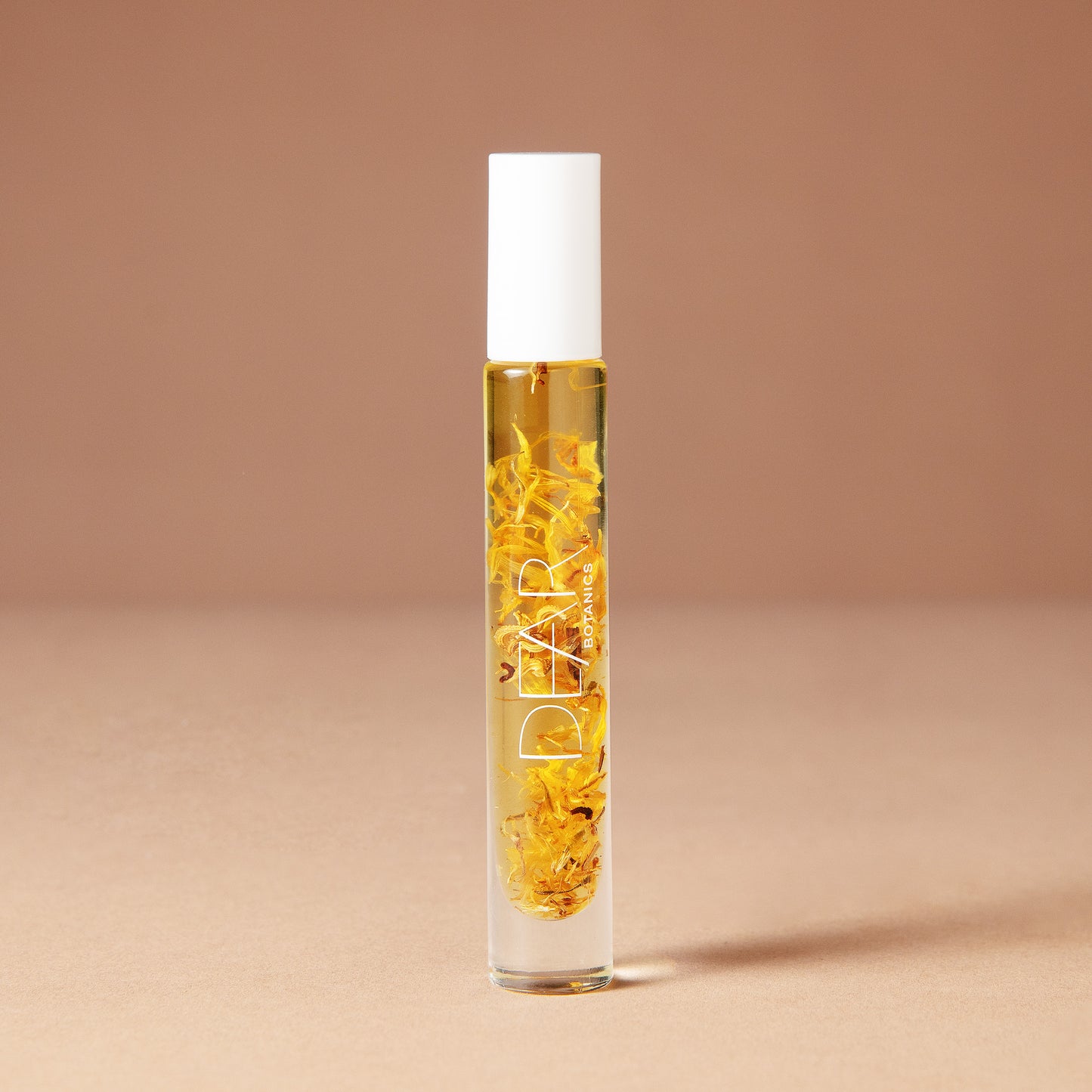 A 10 milliliter clear cylindrical roller bottle filled with pale yellow perfume oil infused with dried flowers featuring a white cap and white vertical text that reads 'DEAR BOTANICS essential oil', set against a solid warm beige background.