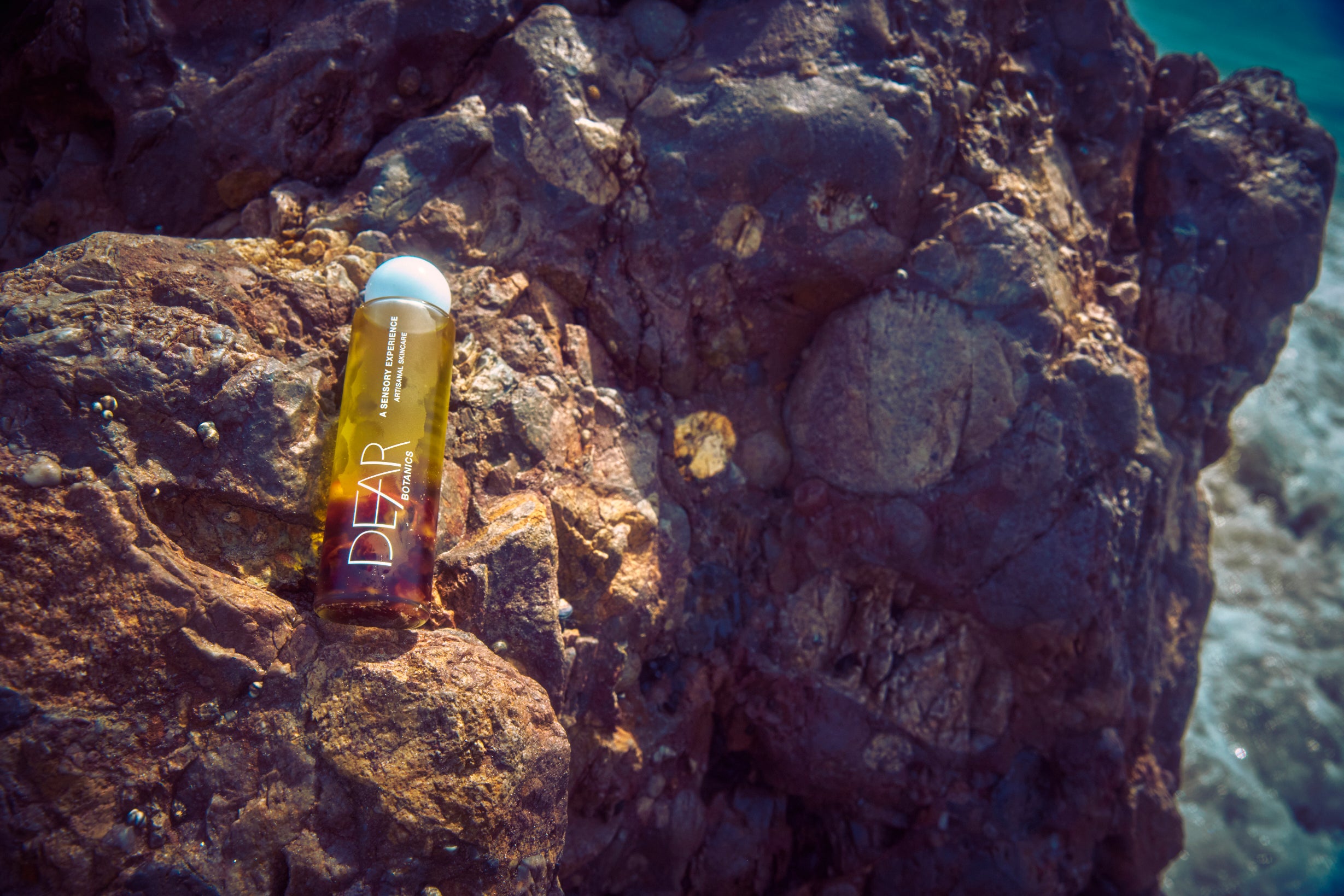 A clear cylindrical bottle containing amber-yellow liquid with a white cap, standing on a large brown rock outdoors. The bottle has white vertical text reading 'DEAR' and smaller text above that appears to say 'MEDITERRANEAN ESSENTIAL OILS'.