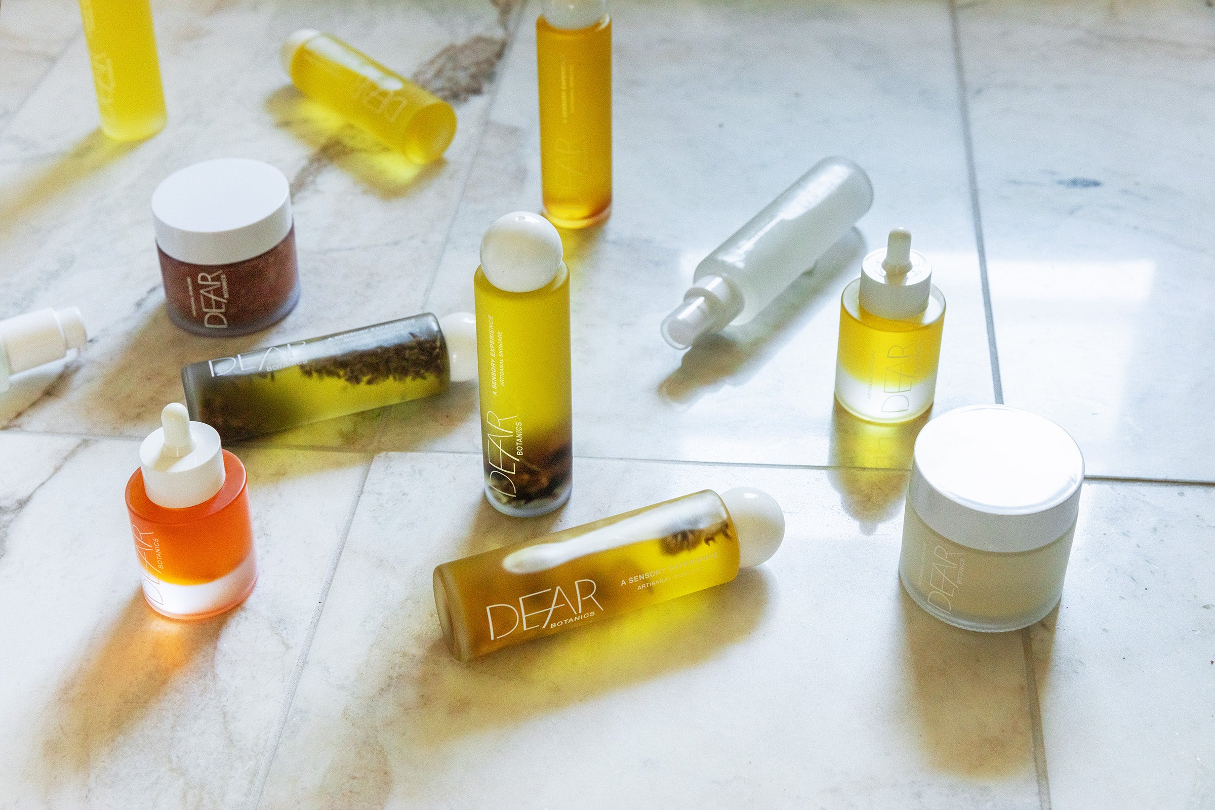 A collection of DEAR brand skincare and cosmetic products, including amber-colored bottles, a white dropper bottle, an orange bottle, a brown jar, and a beige cream jar, arranged on a light marble surface.