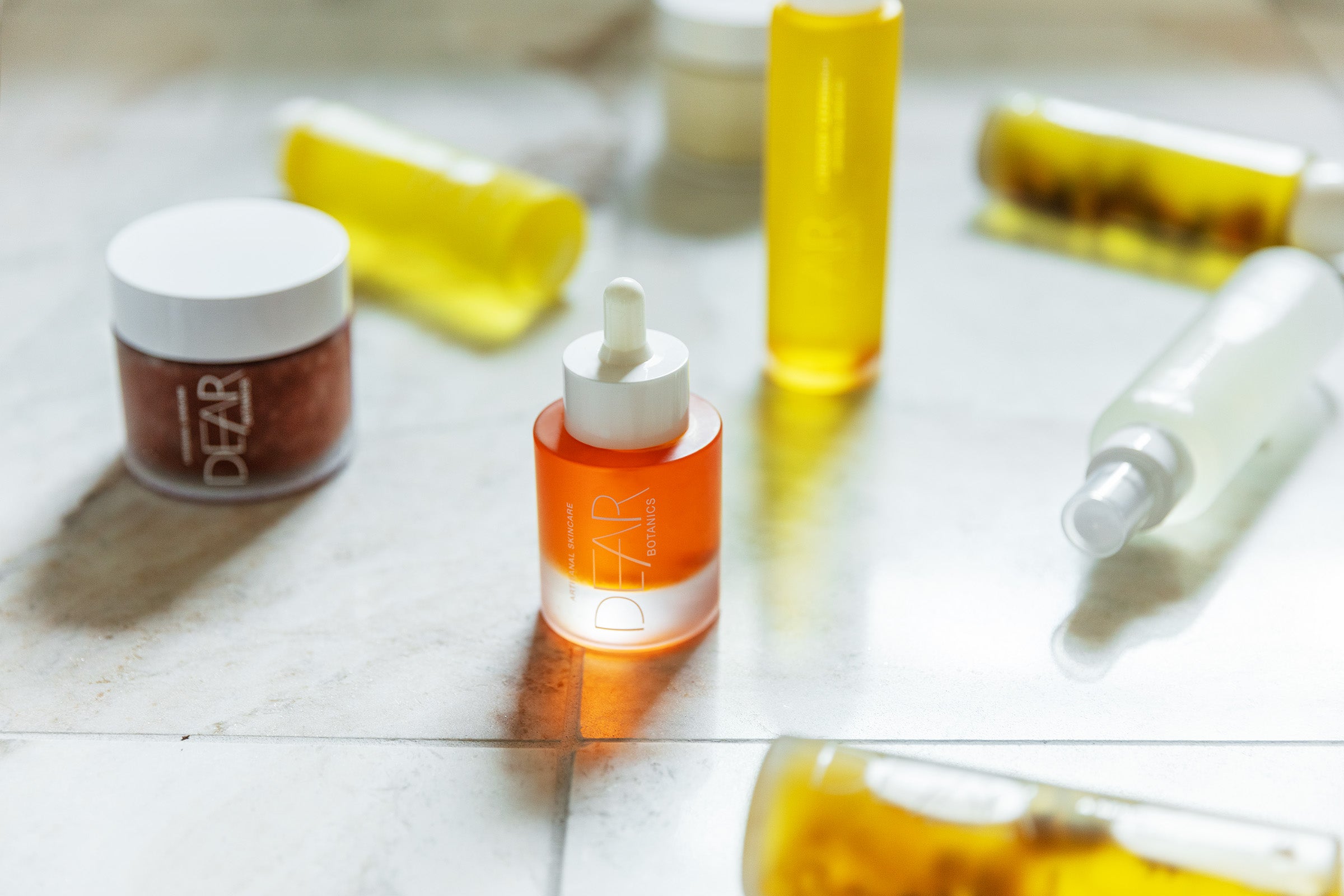A collection of cosmetic bottles and jars from the brand DEAR, including an orange dropper bottle, a brown powder jar, and yellow liquid containers, arranged on a light marble surface with a shallow depth of field.