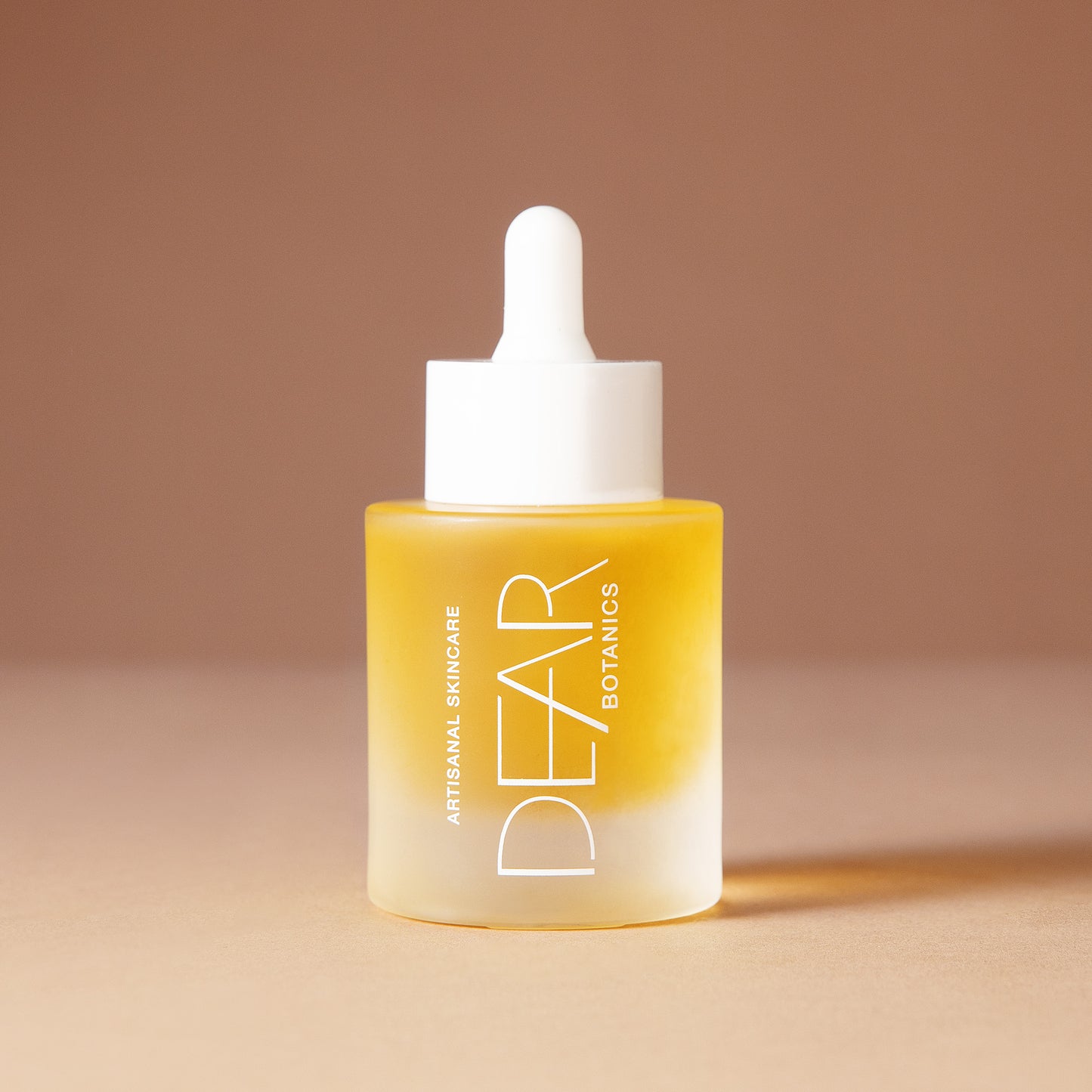 A 50 ml bottle of DEAR BOTANICS Artisanal Skincare face serum with a white dropper cap and a frosted glass bottle showing a golden-yellow liquid inside, set against a warm beige background.