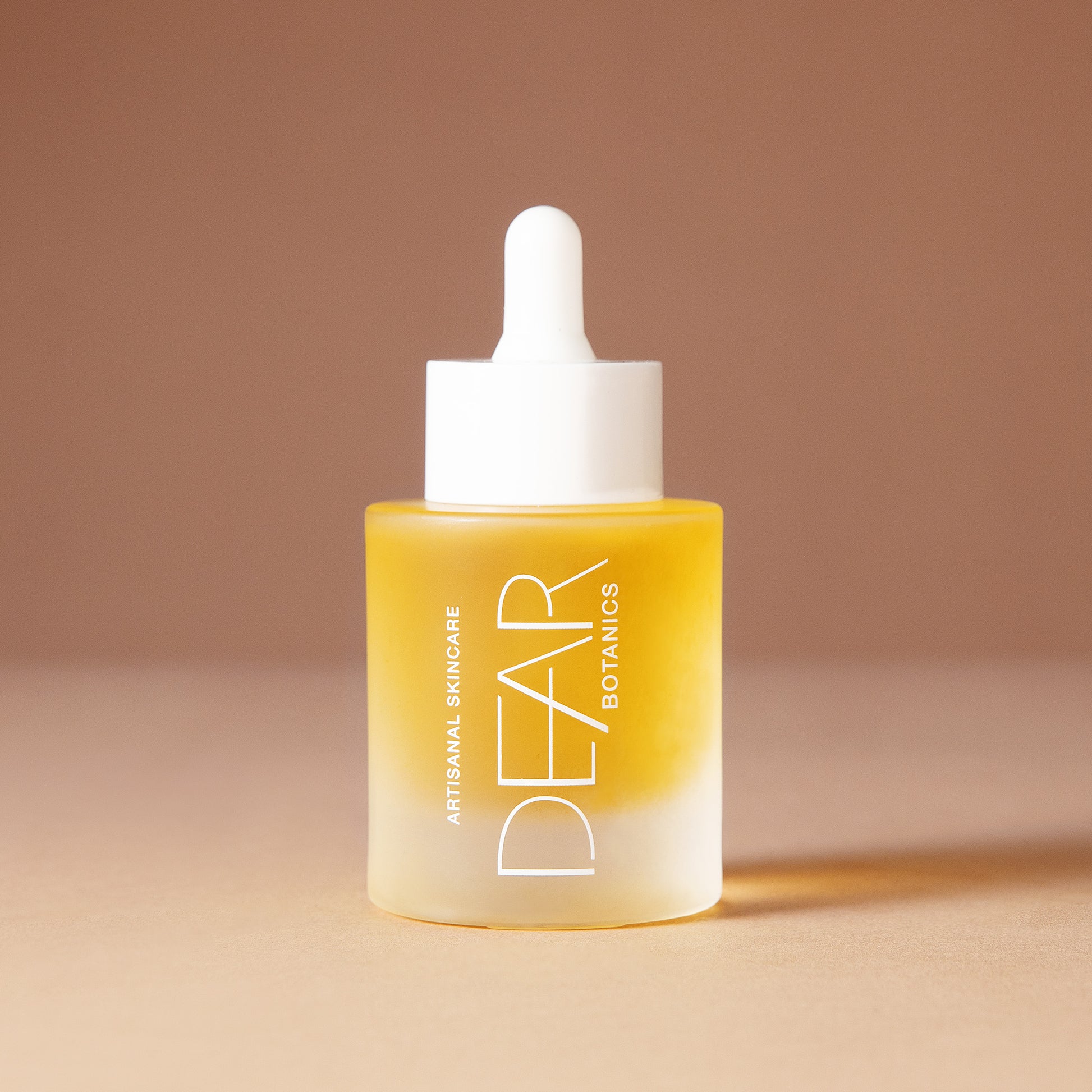 A 50 ml bottle of DEAR BOTANICS Artisanal Skincare face serum with a white dropper cap and a frosted glass bottle showing a golden-yellow liquid inside, set against a warm beige background.
