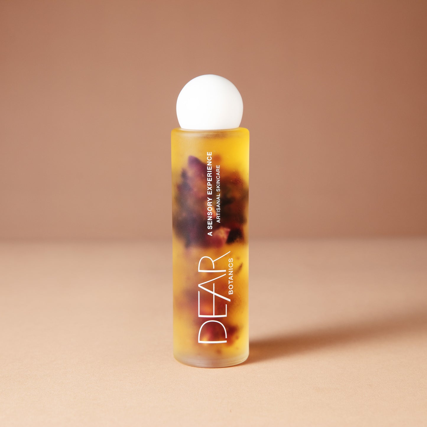 A DEAR BOTANICS 120 ml cosmetic bottle with a white spherical cap, filled with amber-colored liquid containing botanical elements, set against a warm beige background.