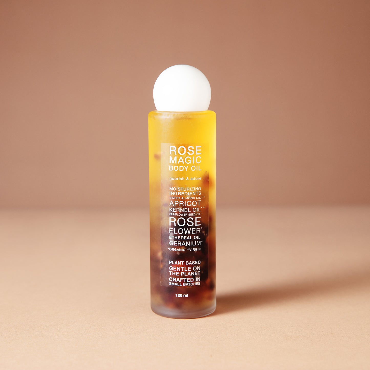 A DEAR BOTANICS 120 ml cosmetic bottle with a white spherical cap, filled with amber-colored liquid containing botanical elements, set against a warm beige background.