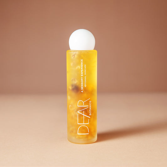 A 120 ml bottle of DEAR BOTANICS essential body oil, featuring a translucent yellow body filled with visible botanical pieces and a white spherical cap, set against a warm beige background.