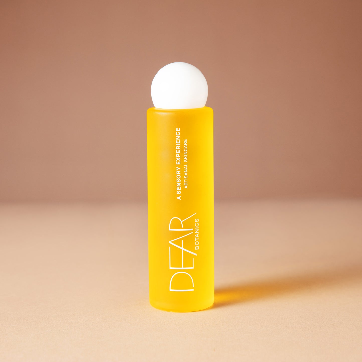 A bright yellow DEAR BOTANICS essential oil 120 ml bottle with a white spherical cap, standing on a beige surface against a warm beige background.