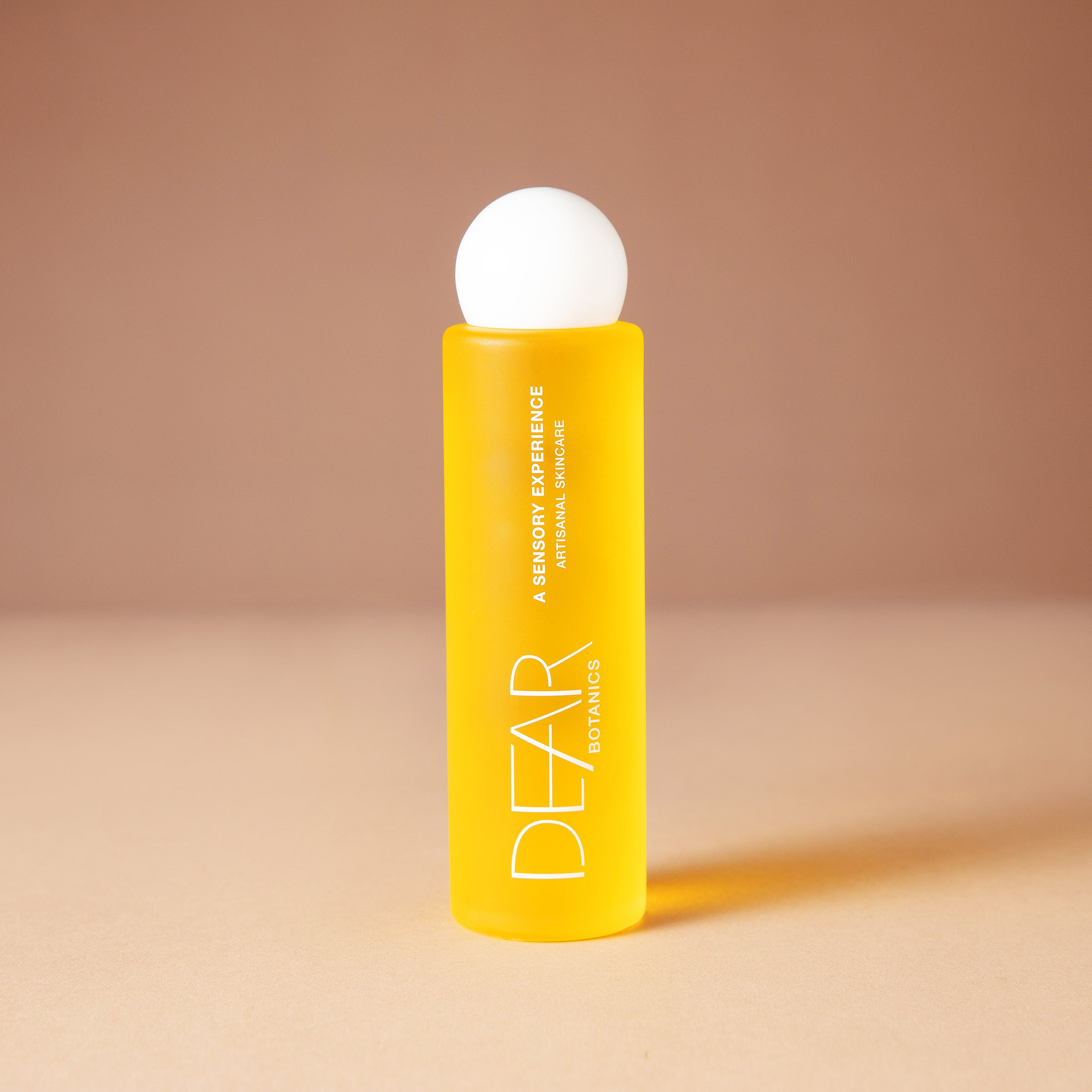 A bright yellow DEAR BOTANICS essential oil 120 ml bottle with a white spherical cap, standing on a beige surface against a warm beige background.