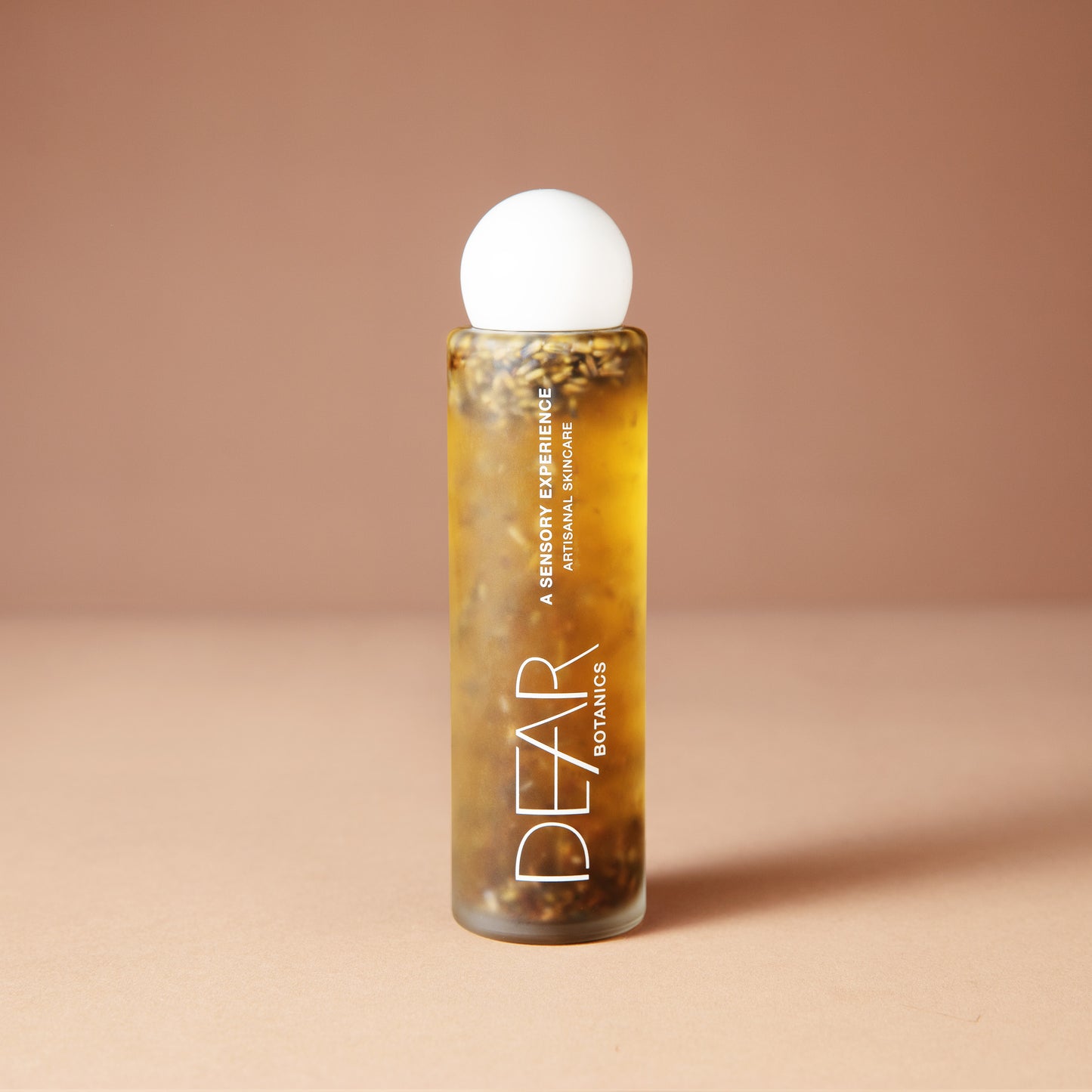 A clear cylindrical 120 ml bottle of DEAR BOTANICS essential oil diffuser liquid with visible botanical pieces inside, topped with a white spherical cap, set against a solid warm beige background.