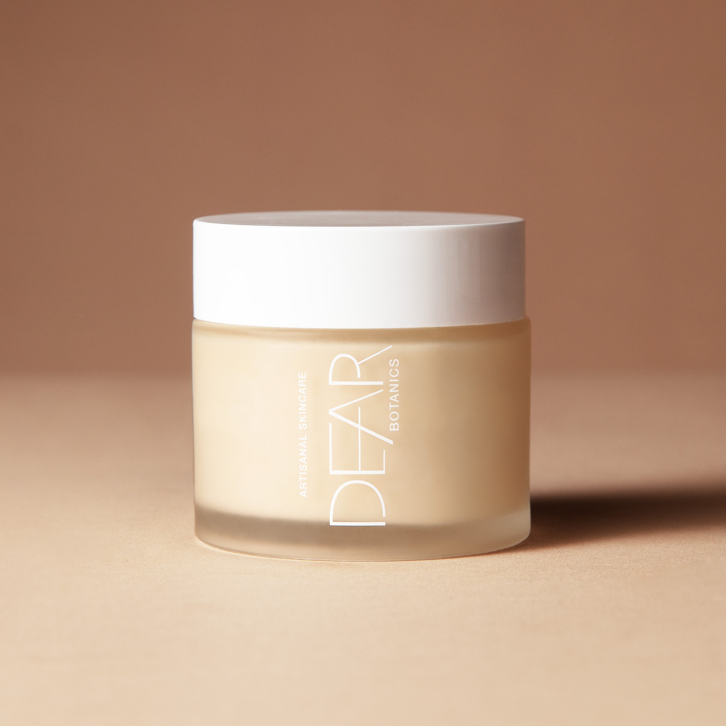 A 150 gram jar of DEAR BOTANICS shea butter cream, featuring a frosted beige glass container and a white lid, set against a warm beige background.
