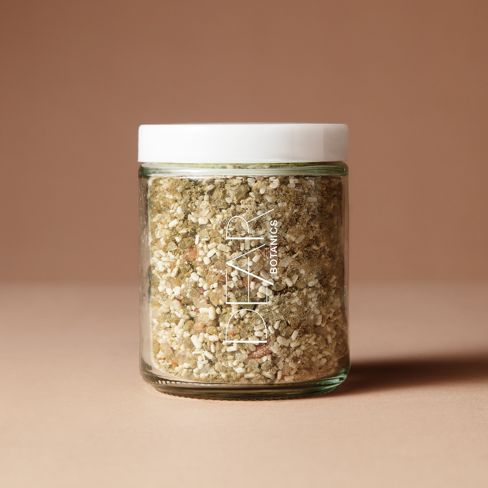 A clear 220 gram glass jar with the white lettering DEAR BOTANICS filled with coarse botanical salt or scrub, sealed with a white lid, standing on a warm beige background. 