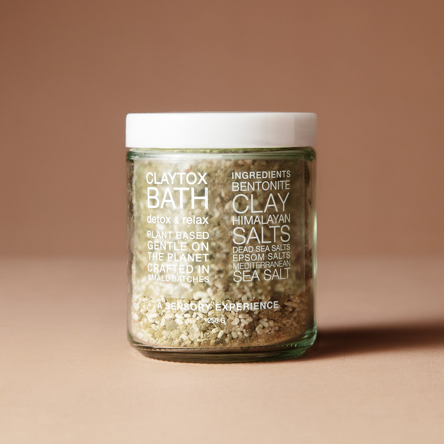 A clear 220 gram glass jar with the ingredients in white lettering filled with coarse botanical salt or scrub, sealed with a white lid, standing on a warm beige background. 