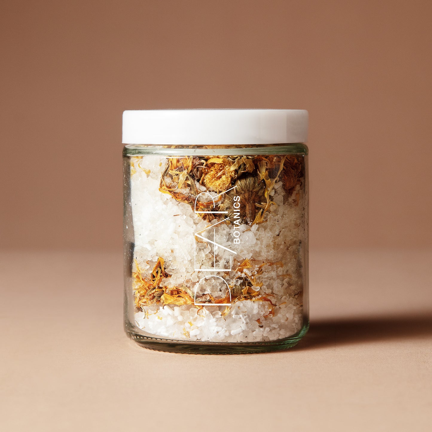 A 220 gram glass jar with white letters DEAR BOTANICS with a white lid containing a mix of dried flowers, salts and crystals on a beige background.