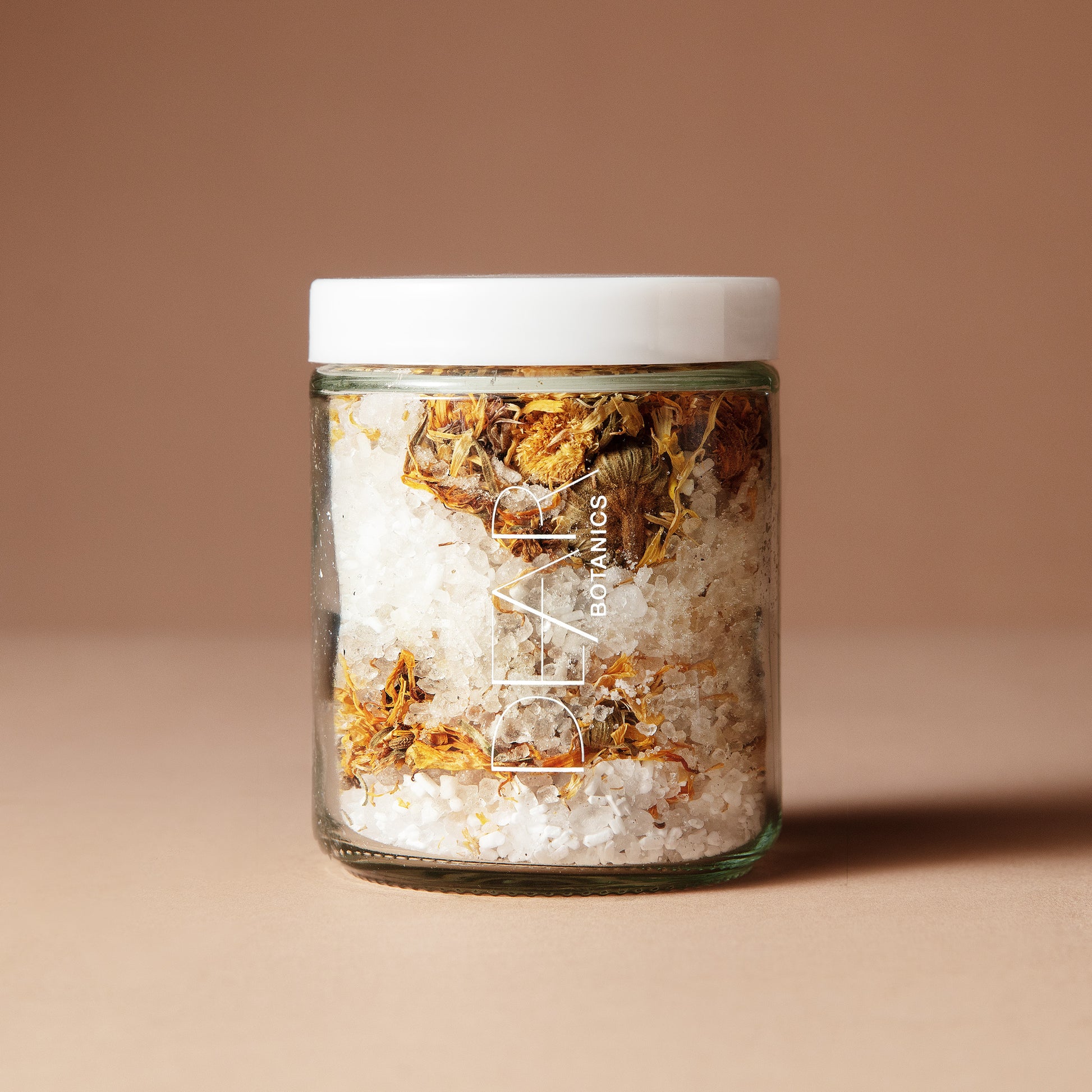 A 220 gram glass jar with white letters DEAR BOTANICS with a white lid containing a mix of dried flowers, salts and crystals on a beige background.