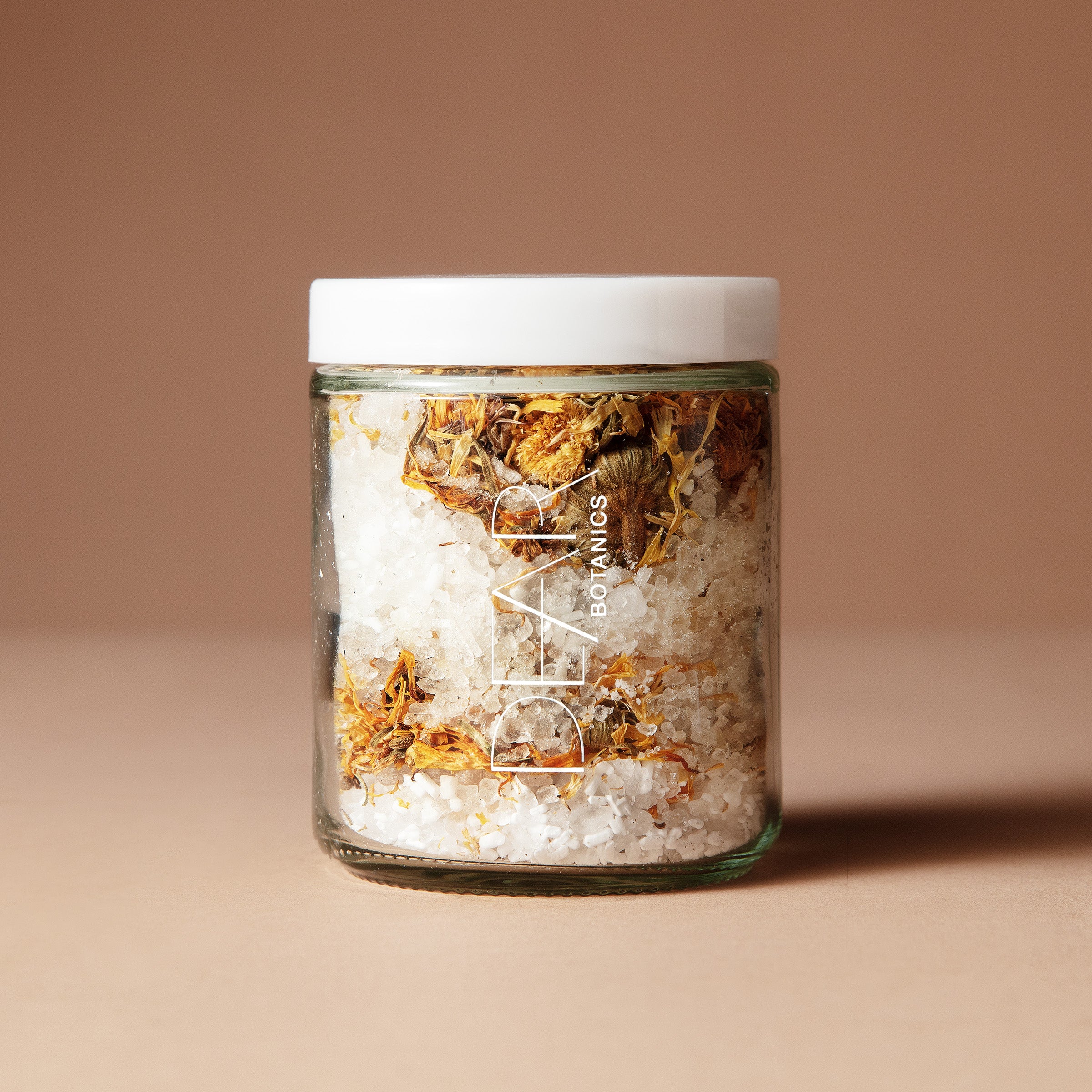A 220 gram glass jar with white letters DEAR BOTANICS with a white lid containing a mix of dried flowers, salts and crystals on a beige background.