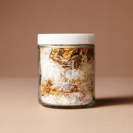 A 220 gram glass jar with white letters DEAR BOTANICS with a white lid containing a mix of dried flowers, salts and crystals on a beige background.