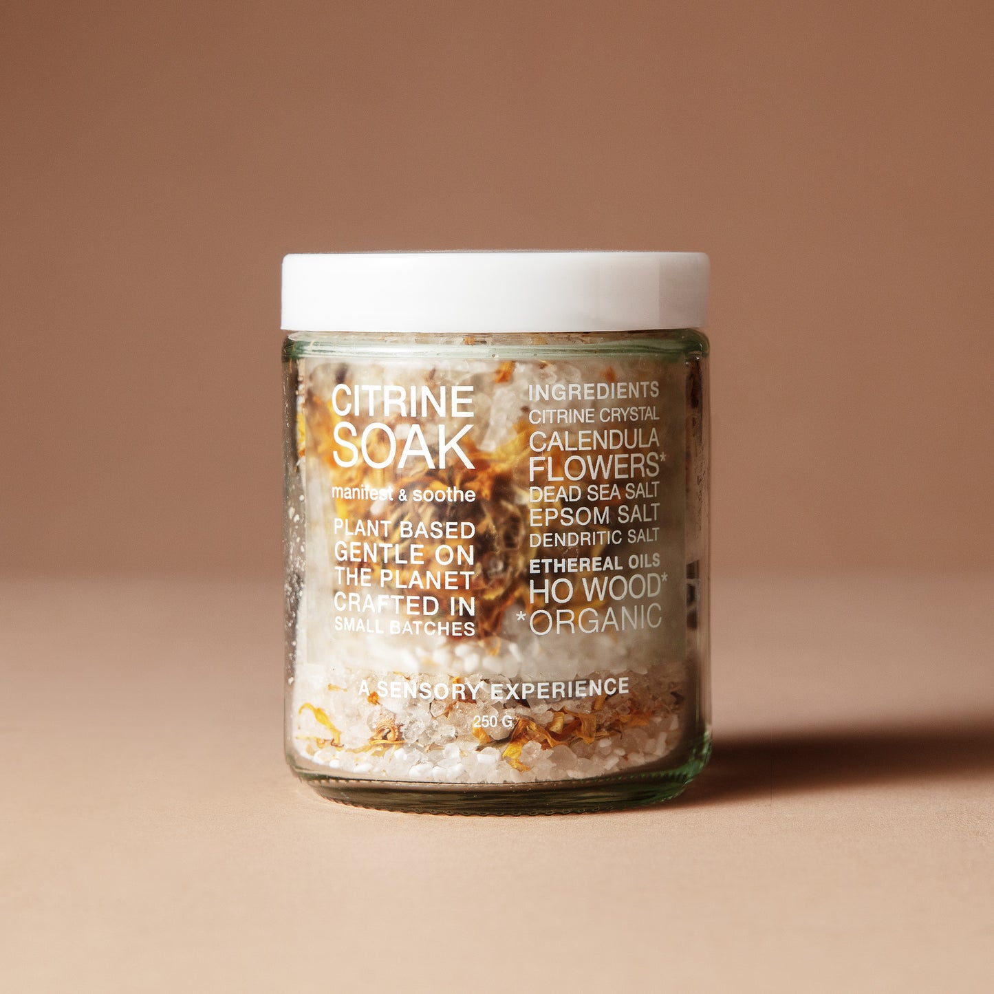 A 220 gram glass jar with white letters of ingredients with a white lid containing a mix of dried flowers, salts and crystals on a beige background.