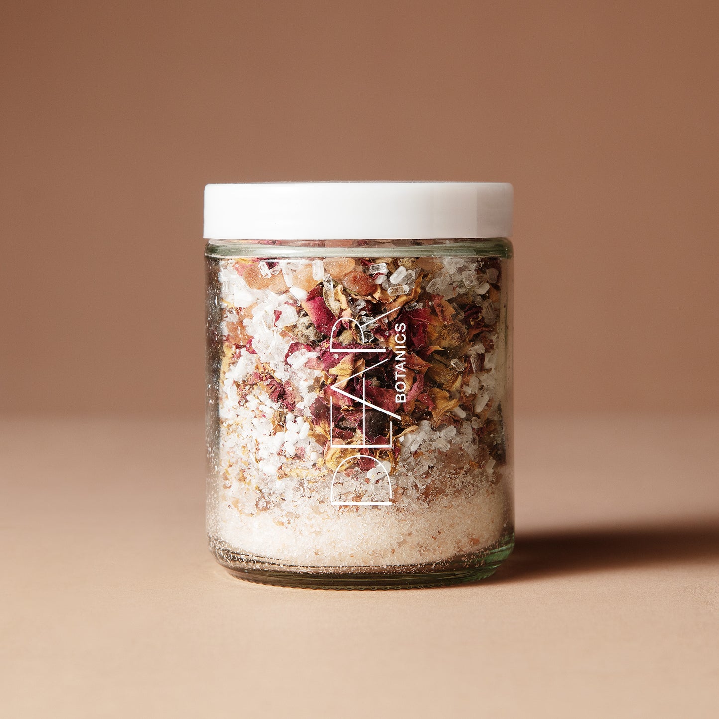 A 220 gram glass jar with white letters DEAR BOTANICS with a white lid containing a mix of dried flowers, salts and crystals on a beige background.