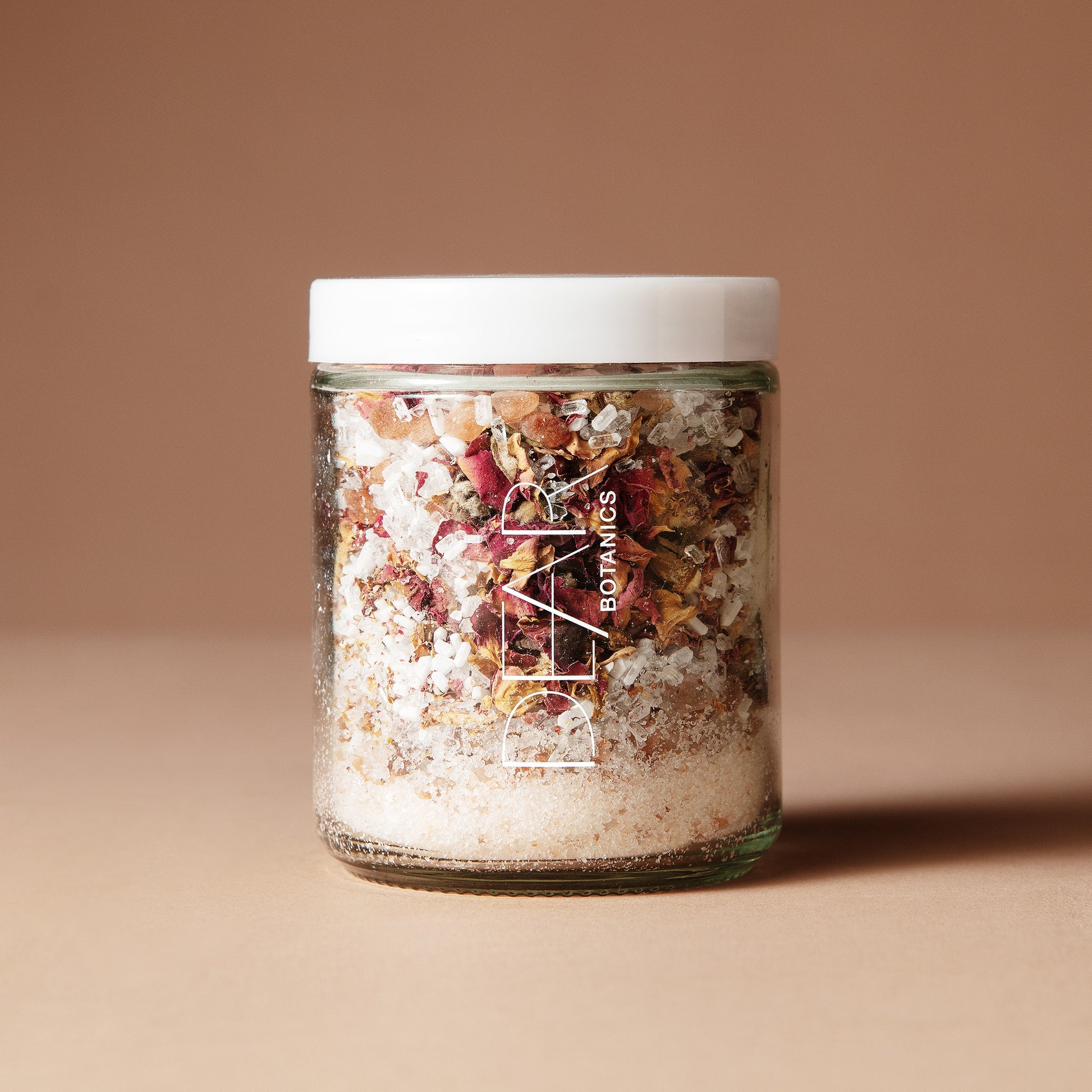 A 220 gram glass jar with white letters DEAR BOTANICS with a white lid containing a mix of dried flowers, salts and crystals on a beige background.