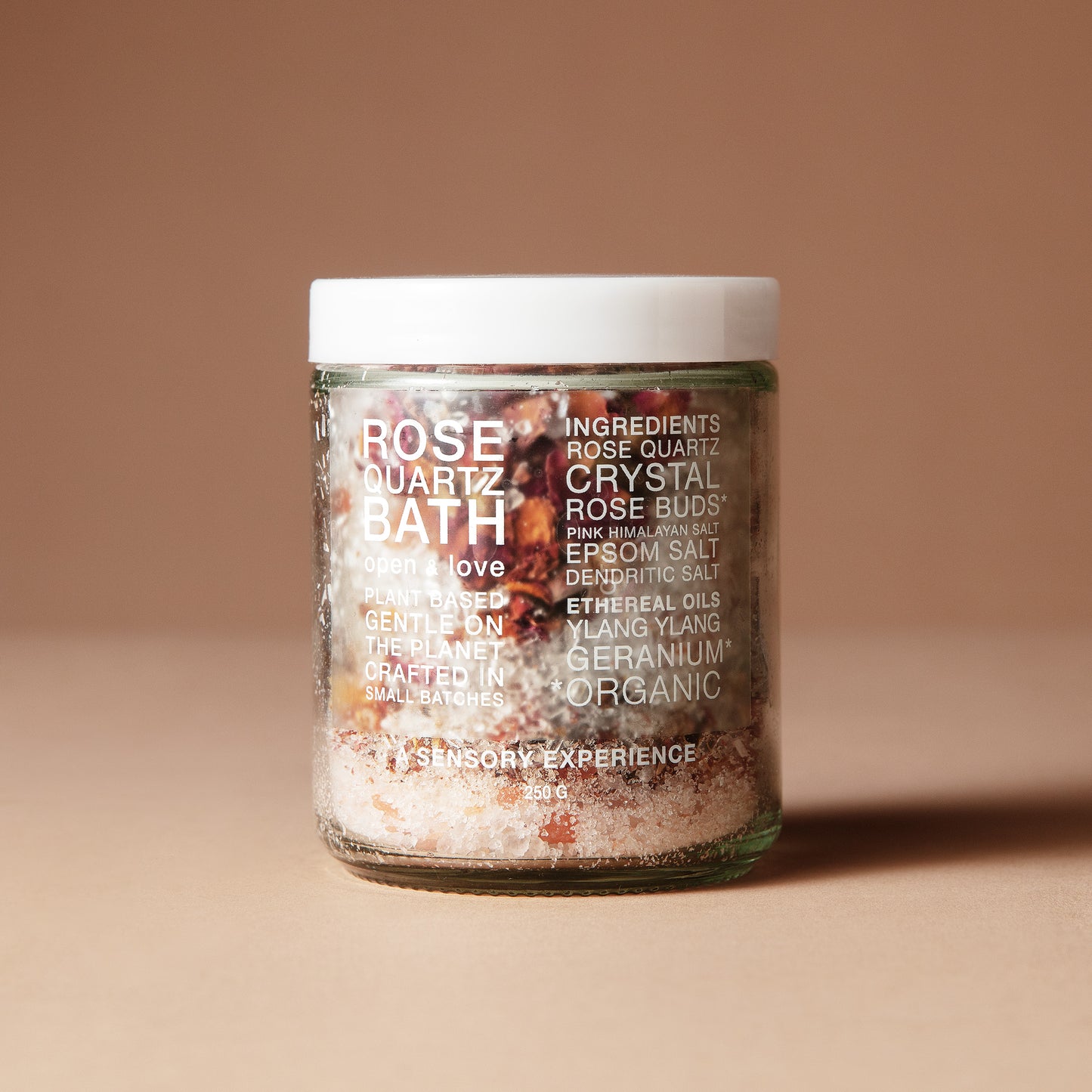 A 220 gram glass jar with white letters of ingredients with a white lid containing a mix of dried flowers, salts and crystals on a beige background.