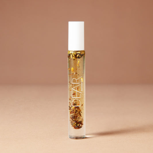 A 10 milliliter clear cylindrical roller bottle filled with pale yellow perfume oil infused with dried flowers featuring a white cap and white vertical text that reads 'DEAR BOTANICS essential oil', set against a solid warm beige background.