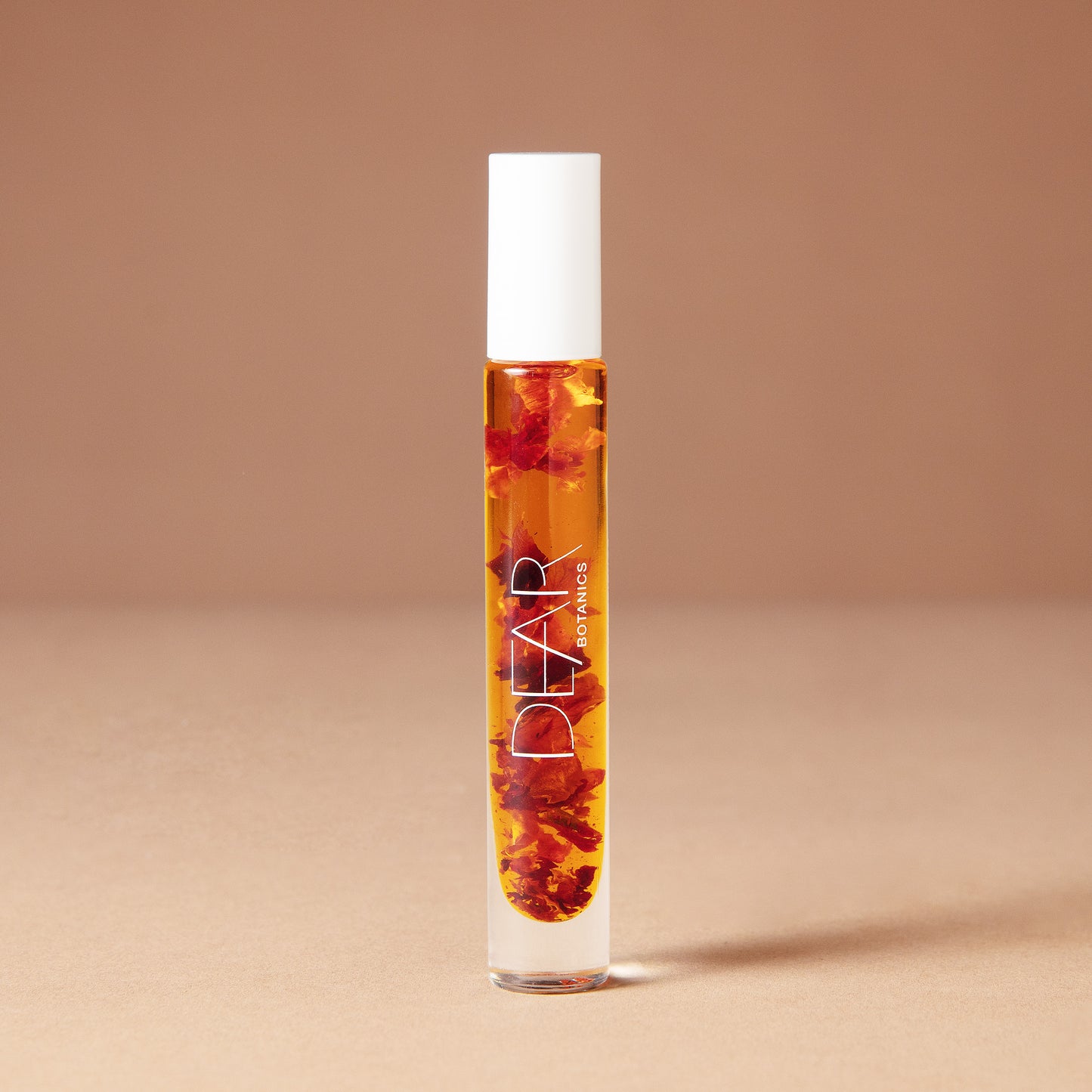 A 10 milliliter clear cylindrical roller bottle filled with pale yellow perfume oil infused with dried flowers featuring a white cap and white vertical text that reads 'DEAR BOTANICS essential oil', set against a solid warm beige background.