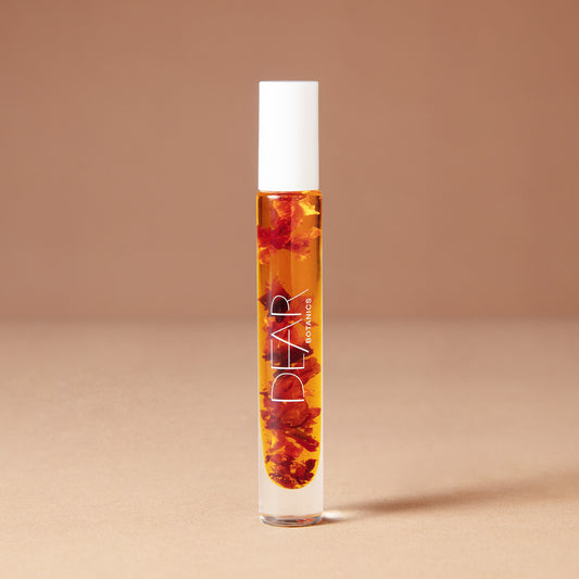 A 10 milliliter clear cylindrical roller bottle filled with pale yellow perfume oil infused with dried flowers featuring a white cap and white vertical text that reads 'DEAR BOTANICS essential oil', set against a solid warm beige background.