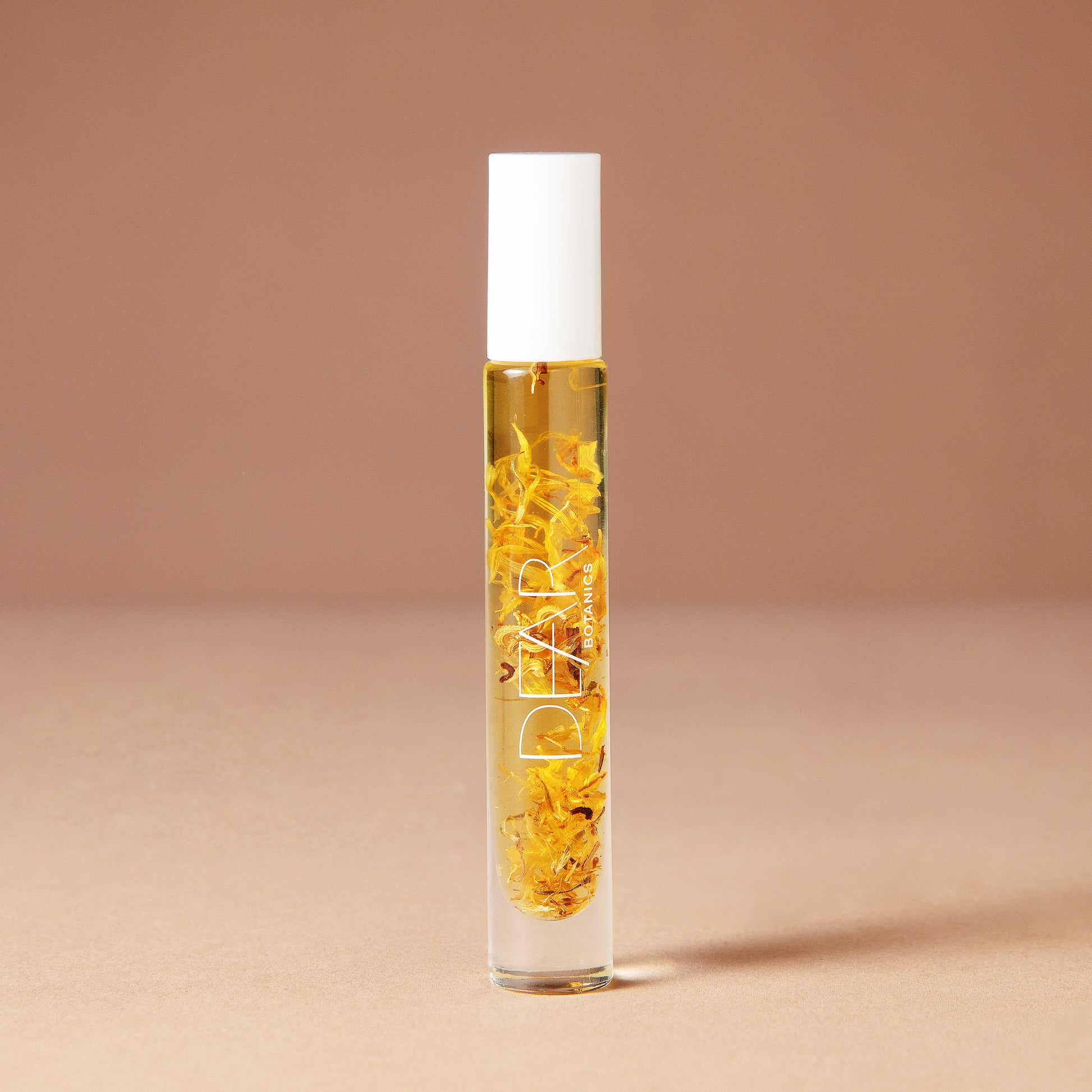A 10 milliliter clear cylindrical roller bottle filled with pale yellow perfume oil infused with dried flowers featuring a white cap and white vertical text that reads 'DEAR BOTANICS essential oil', set against a solid warm beige background.