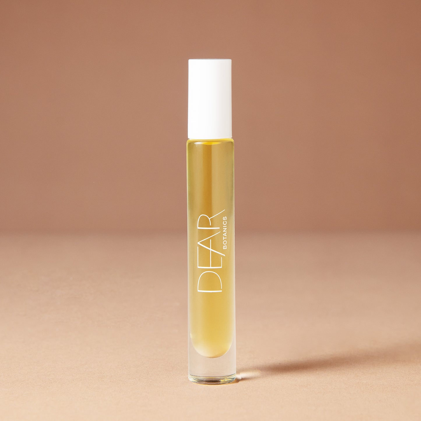A 10 milliliter clear cylindrical roller bottle filled with pale yellow perfume oil, featuring a white cap and white vertical text that reads 'DEAR BOTANICS essential oil', set against a solid warm beige background.
