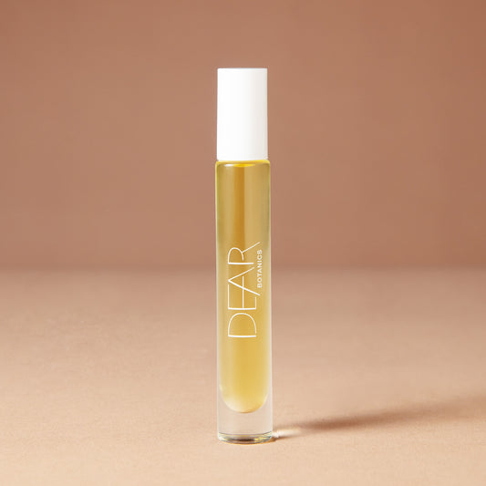 A 10 milliliter clear cylindrical roller bottle filled with pale yellow perfume oil, featuring a white cap and white vertical text that reads 'DEAR BOTANICS essential oil', set against a solid warm beige background.