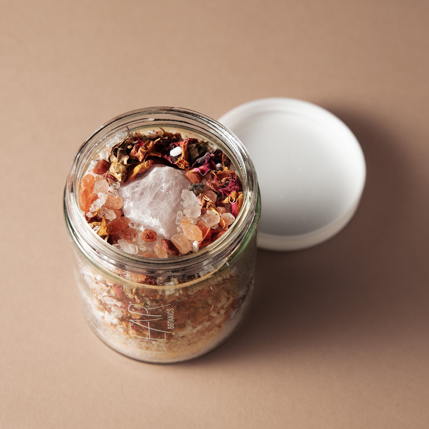 An open clear glass jar filled with a bath or body scrub, topped with dried flowers and crystals, with its white lid resting beside it on a warm beige background.