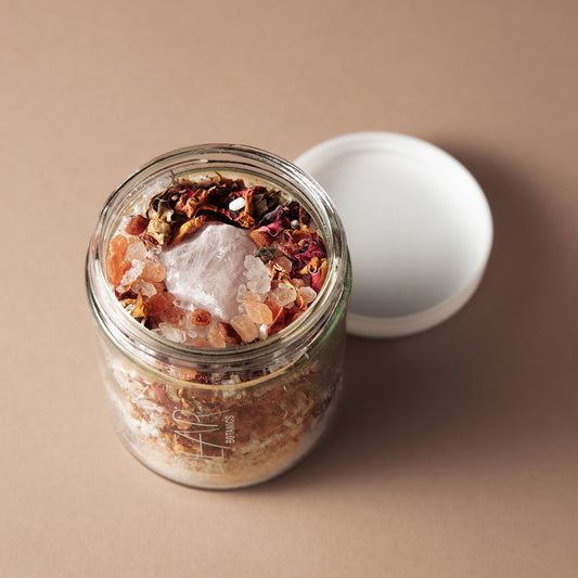 An open clear glass jar filled with a bath or body scrub, topped with dried flowers and crystals, with its white lid resting beside it on a warm beige background.