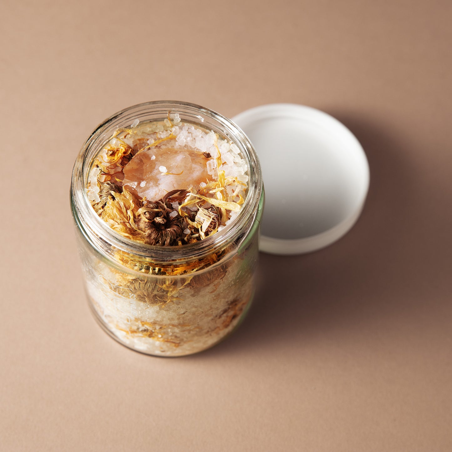 A clear glass jar filled with dried botanicals and coarse salt, shown open with a white lid beside it on a warm beige background.