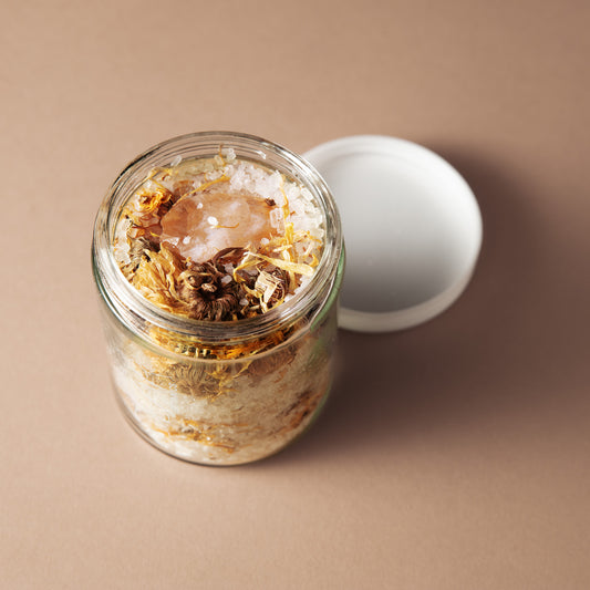 A clear glass jar filled with dried botanicals and coarse salt, shown open with a white lid beside it on a warm beige background.