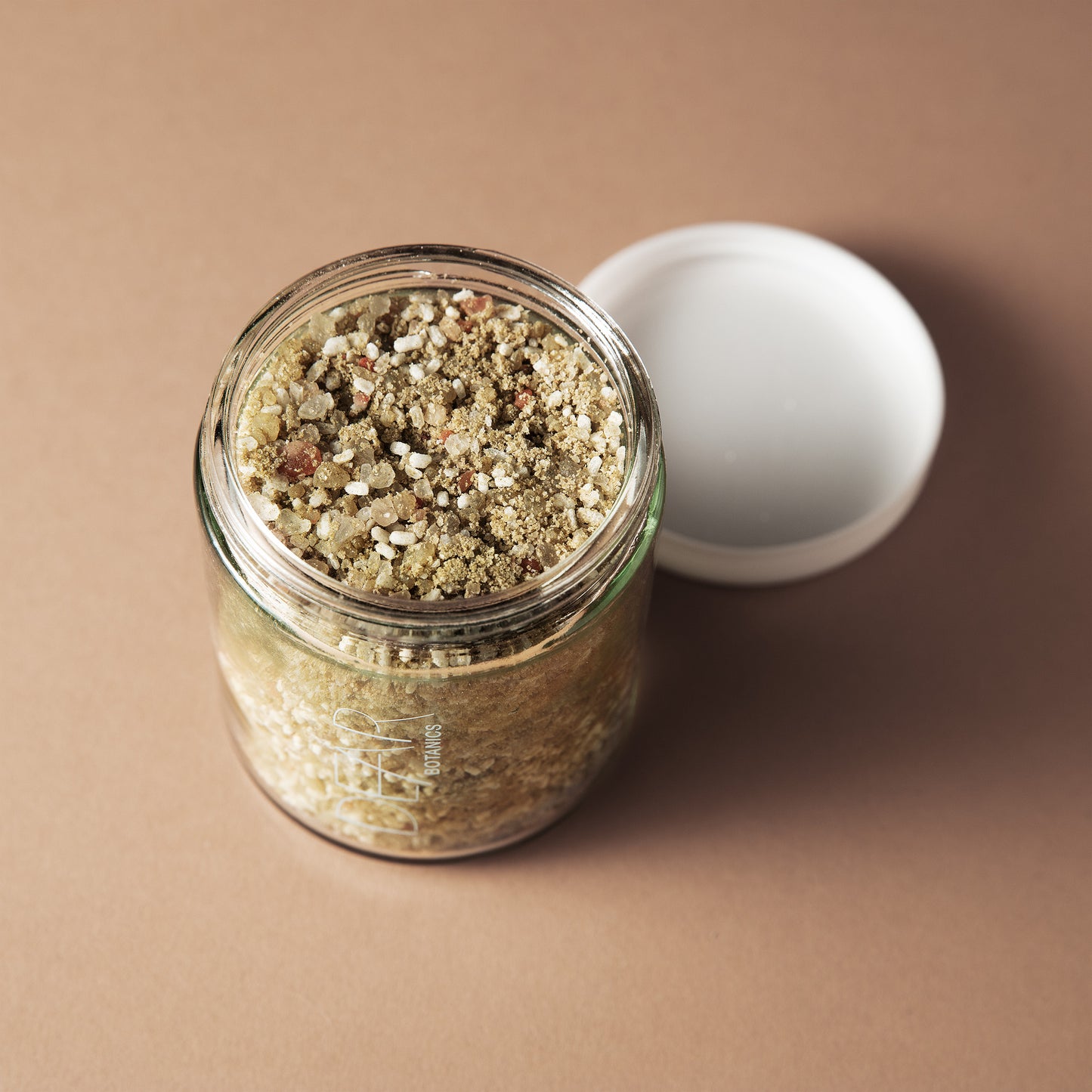 An open clear glass jar filled with a granular beige and olive-brown mixture, shown on a warm tan background with a white lid resting beside it.